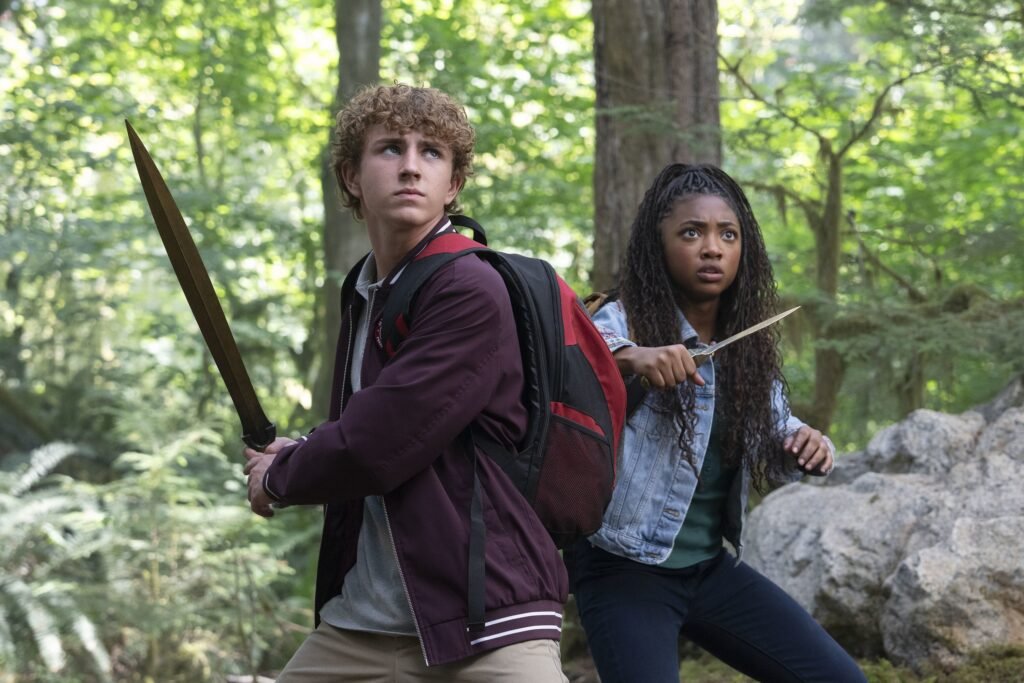 Walker Scobell (Percy Jackson) e Leah Jeffries (Annabeth Chase) em pose de combate na floresta. A imagem destaca a química e a prontidão dos jovens protagonistas na adaptação.